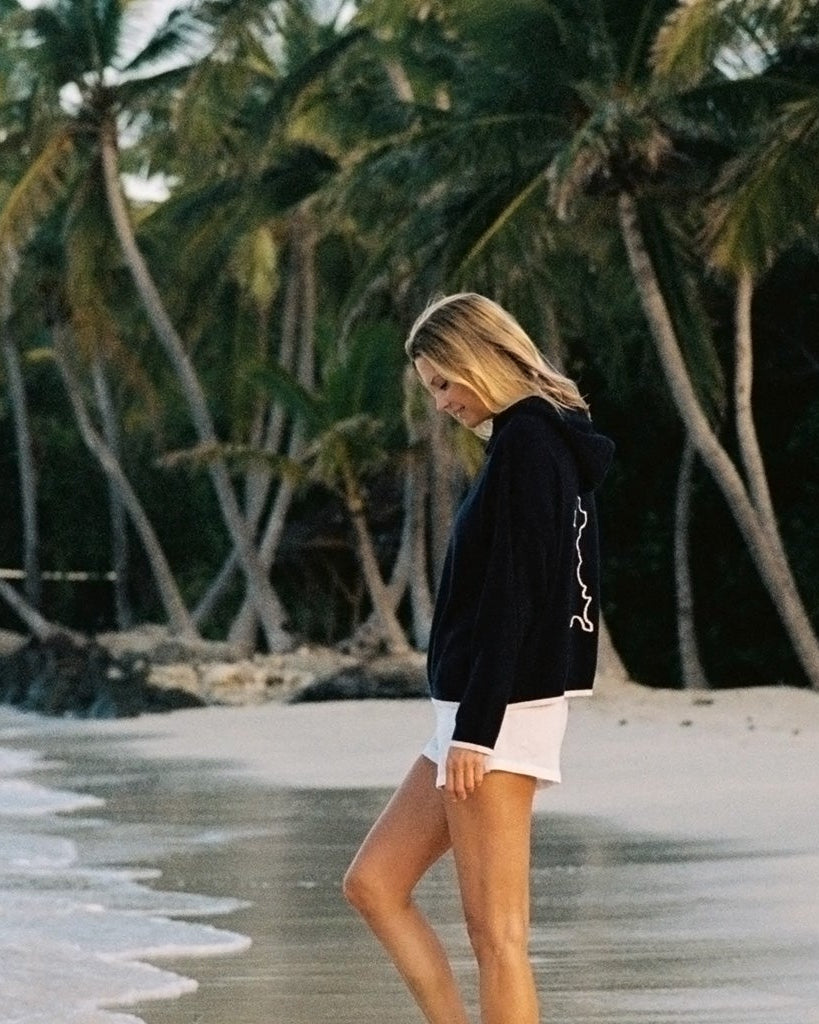 Women’s New Arrivals - Malin modeling a luxury navy cashmere hoodie on lagoon bay, Mustique Island
