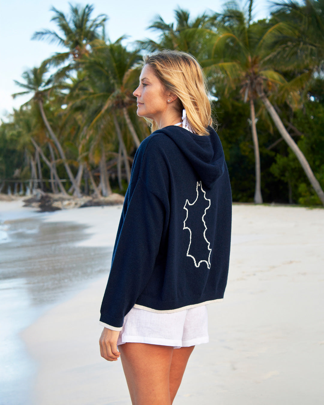 Woman wearing a navy cashmere hoodie with a white logo on a beach with palm trees in the background