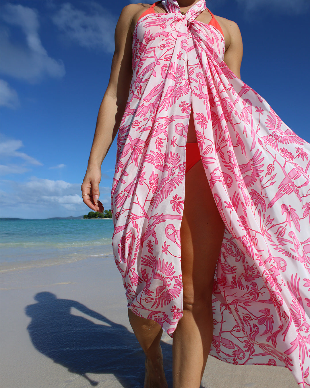 Woman wearing Pink House pure silk crepe-de-Chine sarong in Parrot Pink tied as beach cover-up, walking along Mustique shoreline.