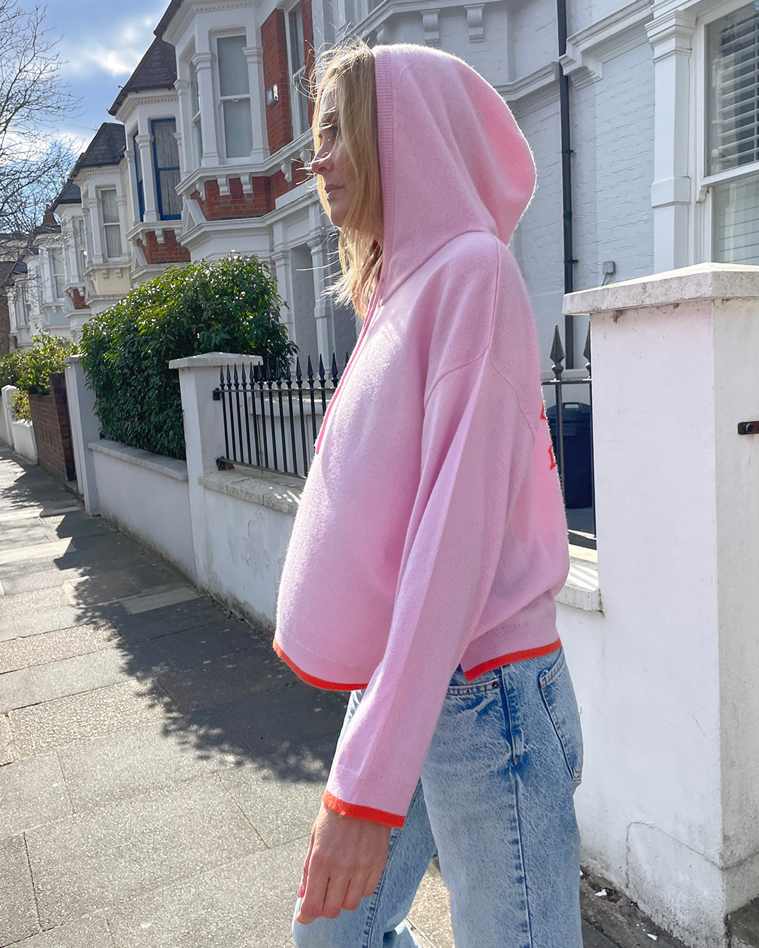 Person wearing a pink cashmere hoodie and blue jeans standing on a sidewalk with residential buildings in the background.