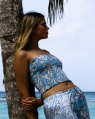 Model leans on palm tree at Lagoon Bay Mustique wearing blue parrot print silk bandeau top and matching silk satin PJs