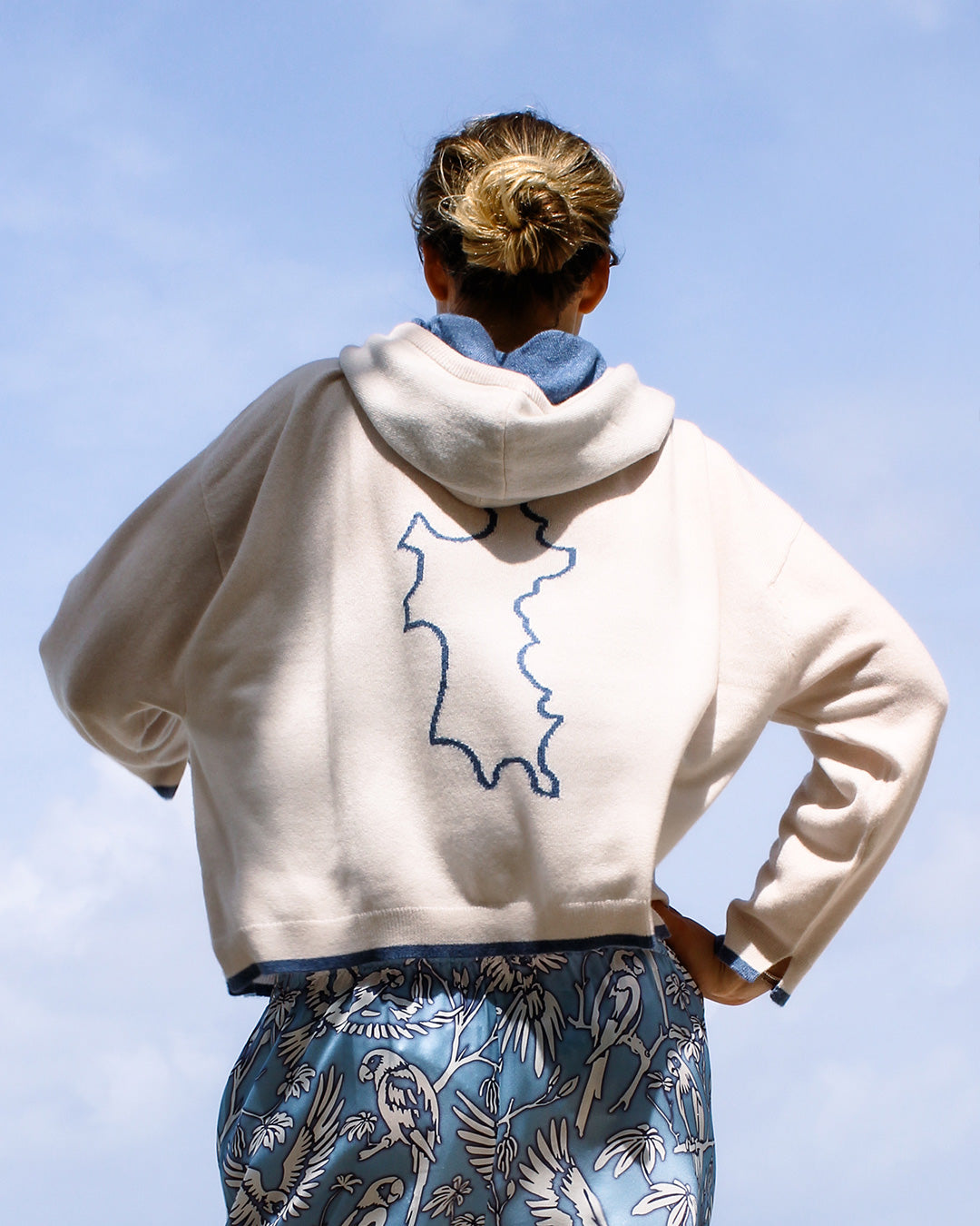 Person wearing a white cashmere hoodie with a blue design on the back against a clear blue sky.