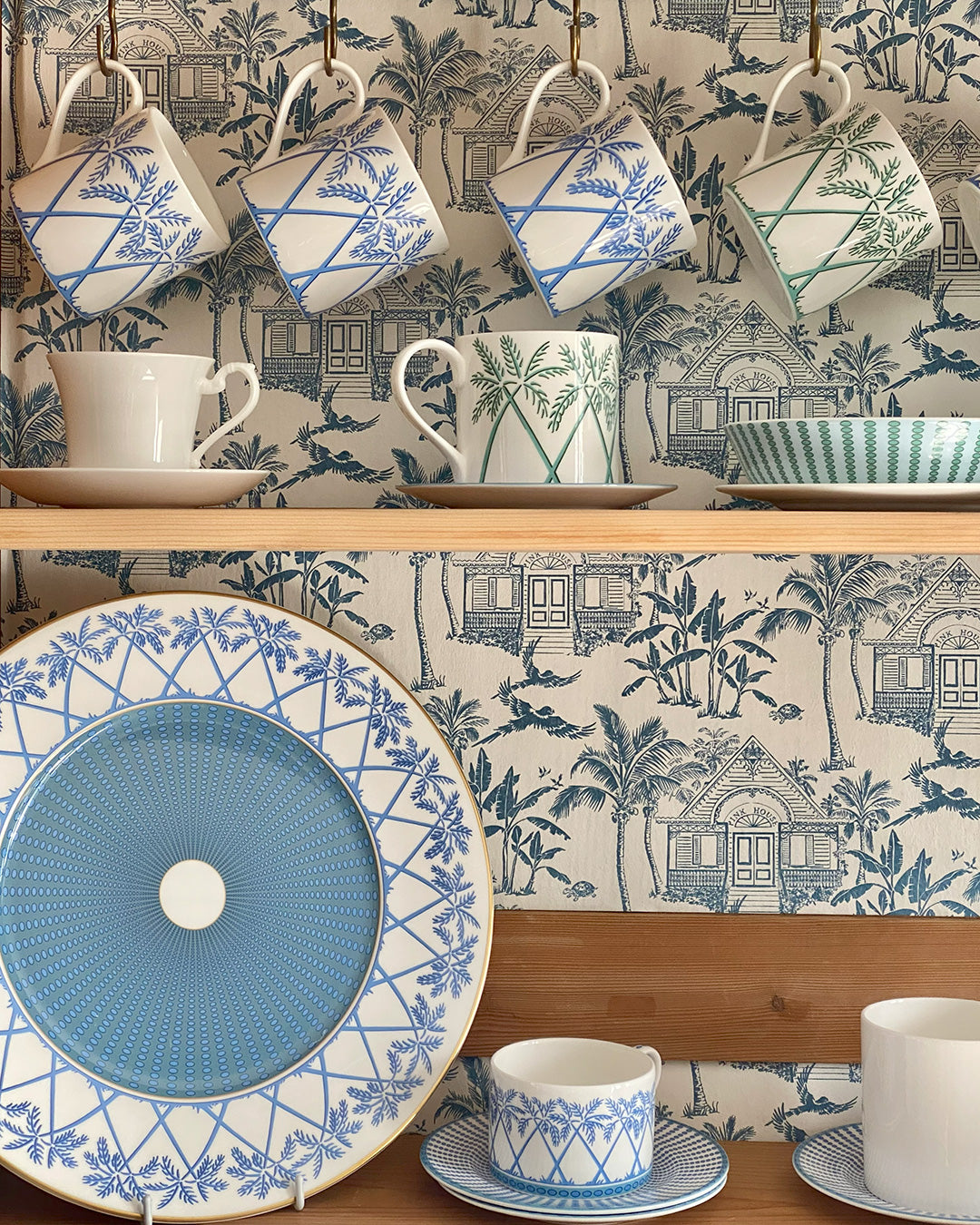 Fine bone porcelain china mugs and charger plate in Palms Azure Blue design, styled in a dresser cupboard – Pink House Mustique luxury tableware.