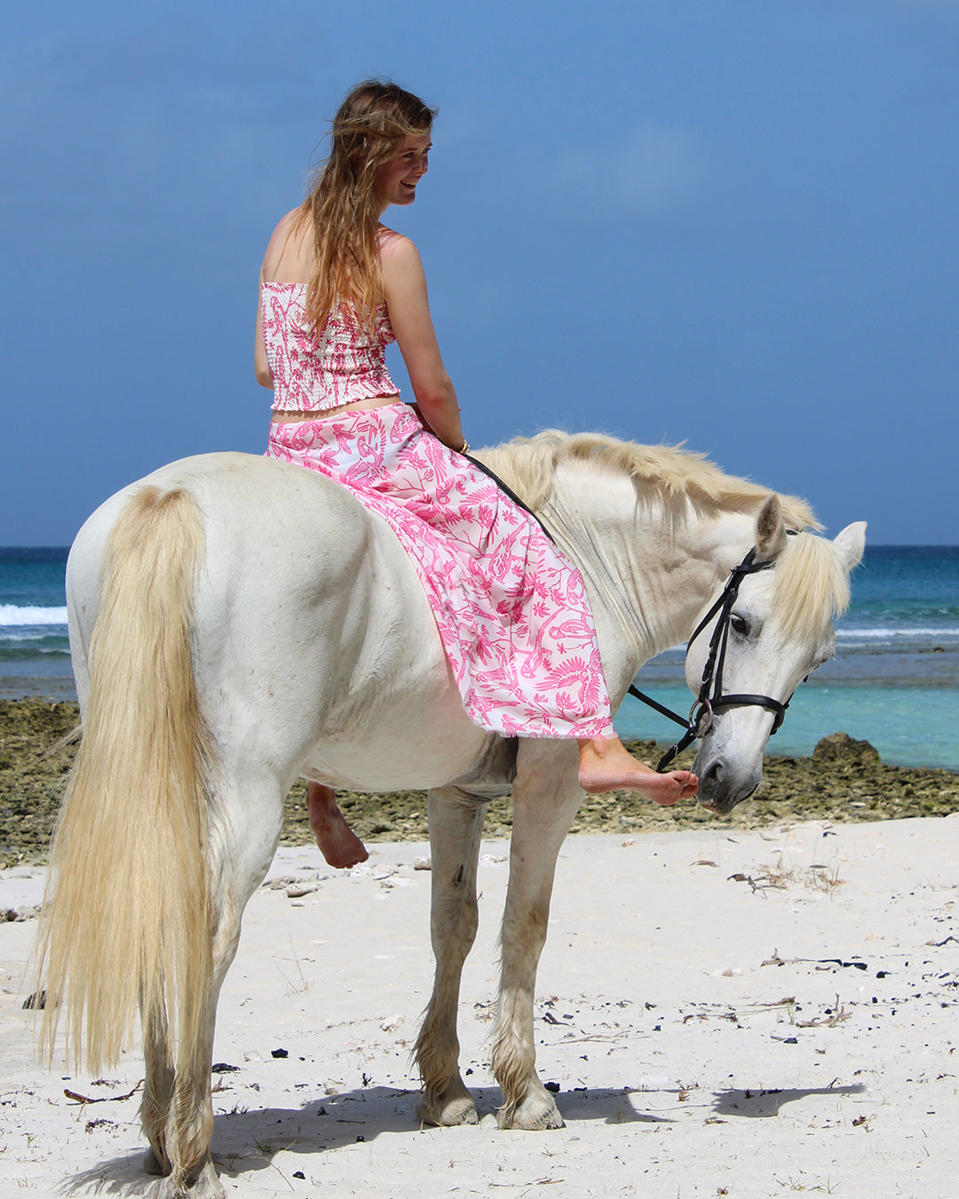 Model in Parrot Pink silk pareo and bandeau set riding white horse on Mustique beach – feminine island resort look
