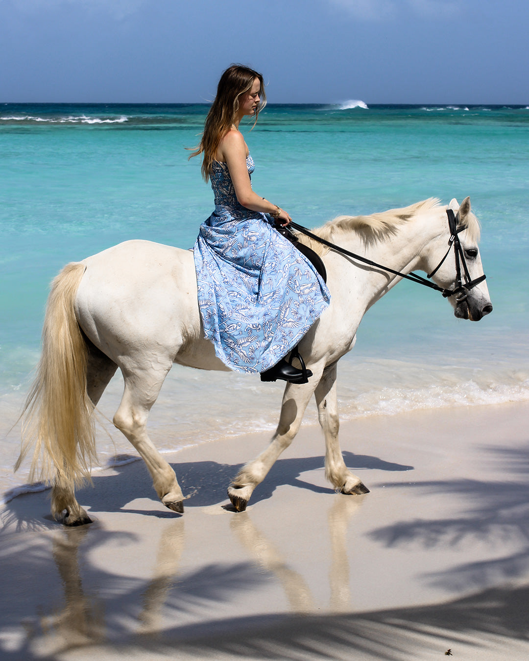 Model wearing pure silk sarong in Parrot Blue riding white horse on Mustique beach, luxury island resortwear look