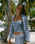 Wearing full silk co-ord in parrot blue, model poses on beach at Lagoon Bay Mustique in bandeau top and matching PJ set