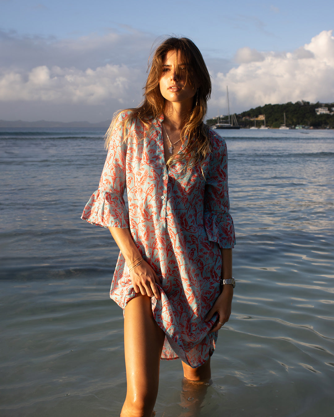 Lottie wearing Pink House Linen Decima Dress in Parrot Coral/Turquoise standing in tropical ocean water – luxury resort wear with parrot print and gathered sleeves.