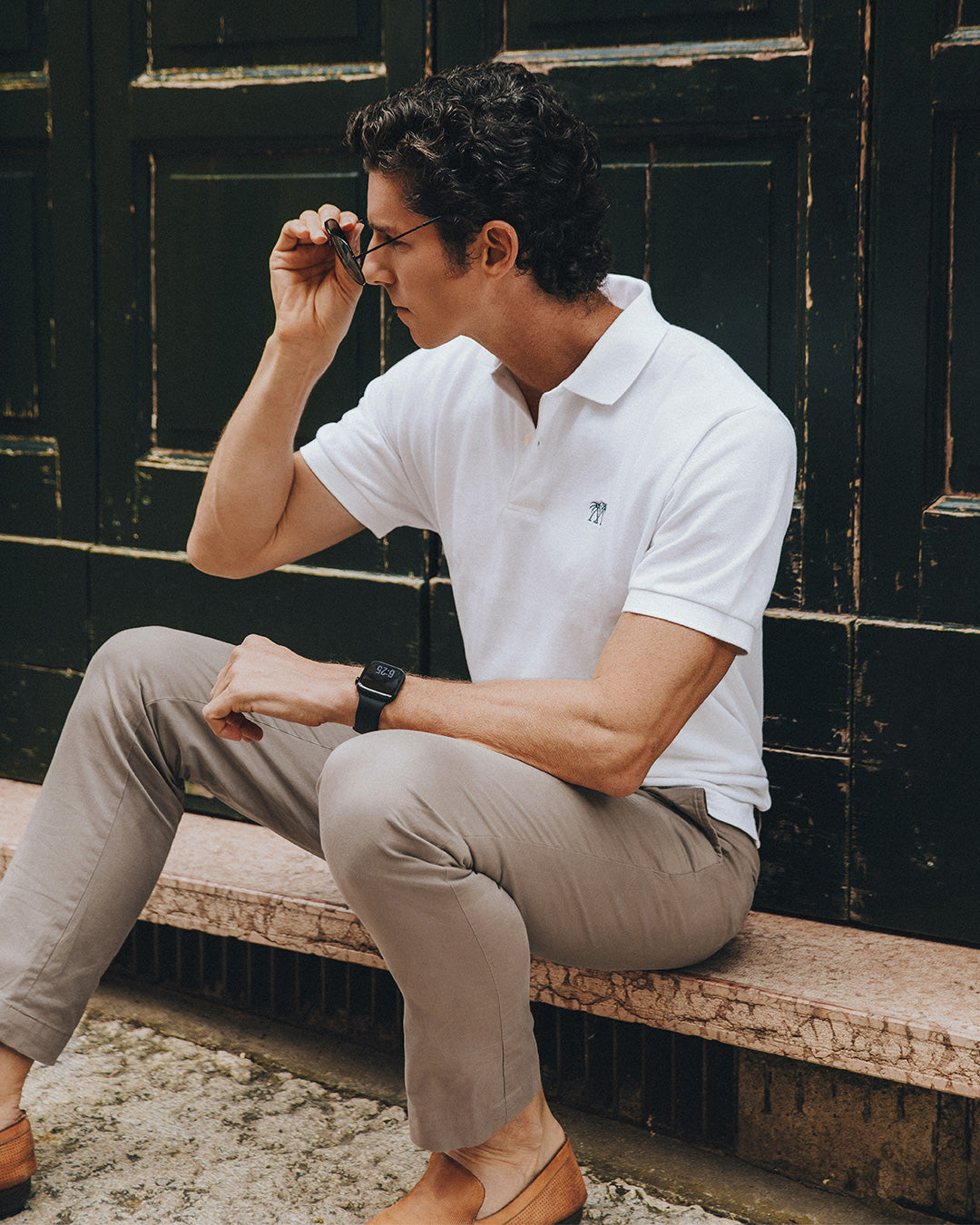 Close-up of a men's white cotton polo shirt with embroidered palm logo in a Mediterranean lifestyle setting.