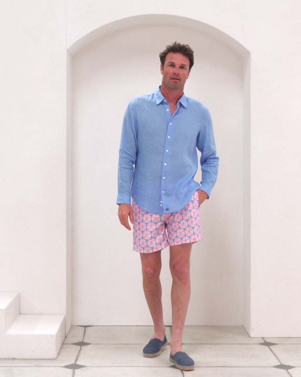 Mens recycled swim shorts in pink and blue Sand Dollar print by Pink House Mustique worn in studio by Joss paired with French Blue linen shirt