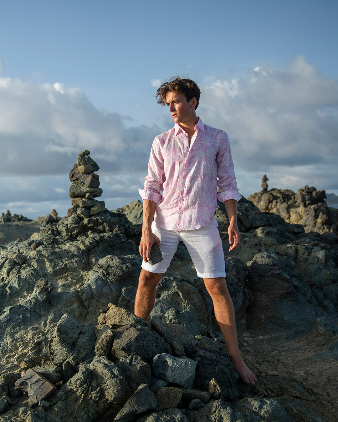 Men’s Agave pink linen shirt by Pink House Mustique, styled with white linen shorts on coastal rocks.