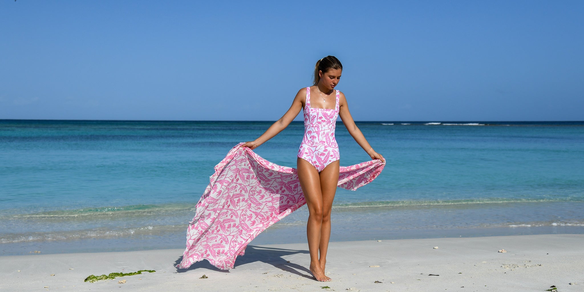 Luxury women’s swimwear collection by Pink House Mustique – model on Lagoon Beach wearing Parrot Pink one-piece swimsuit with matching silk cover-up.