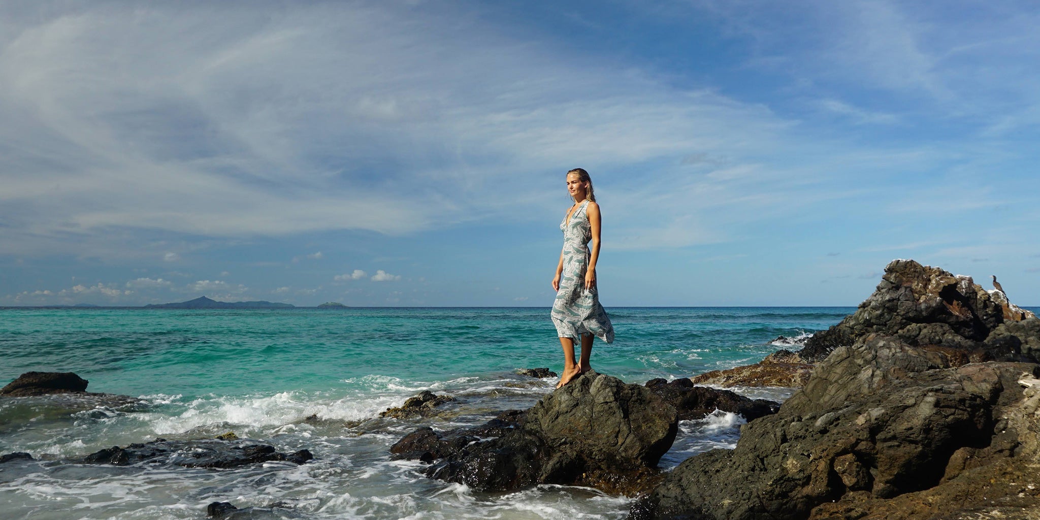 Model wearing Whale print silk Jemima dress on the rocks beside the turquoise sea at Gelliceaux Beach, Mustique
