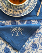Blue linen napkin embroidered with crossed palms on pomegranate print tablecloth, luxury dining linens by Pink House Mustique.