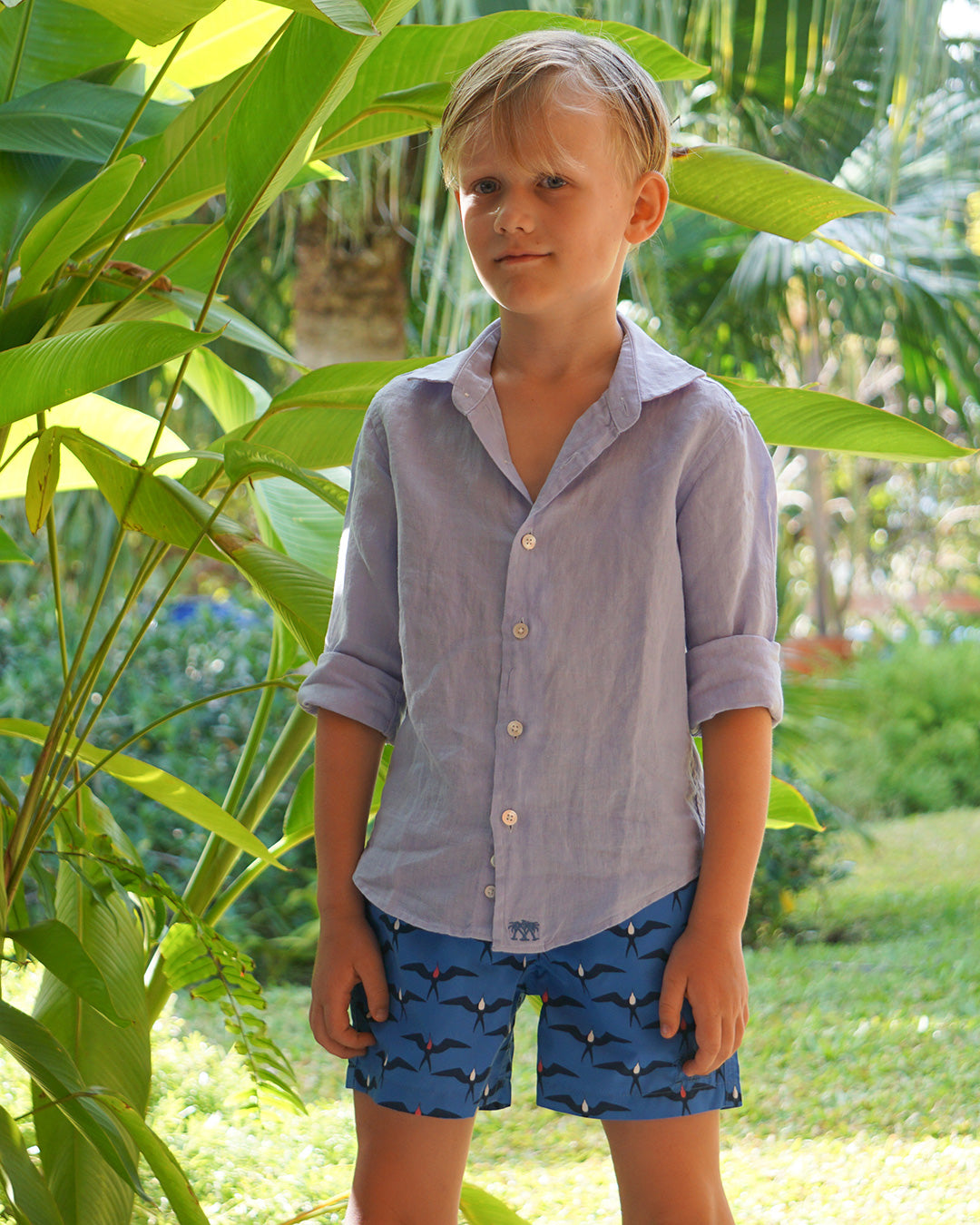 Boy wearing Frigate Bird Blue swim shorts and azul blue linen shirt in tropical garden