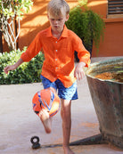Boy wearing Frigate Bird Blue swim shorts and orange linen shirt playing football