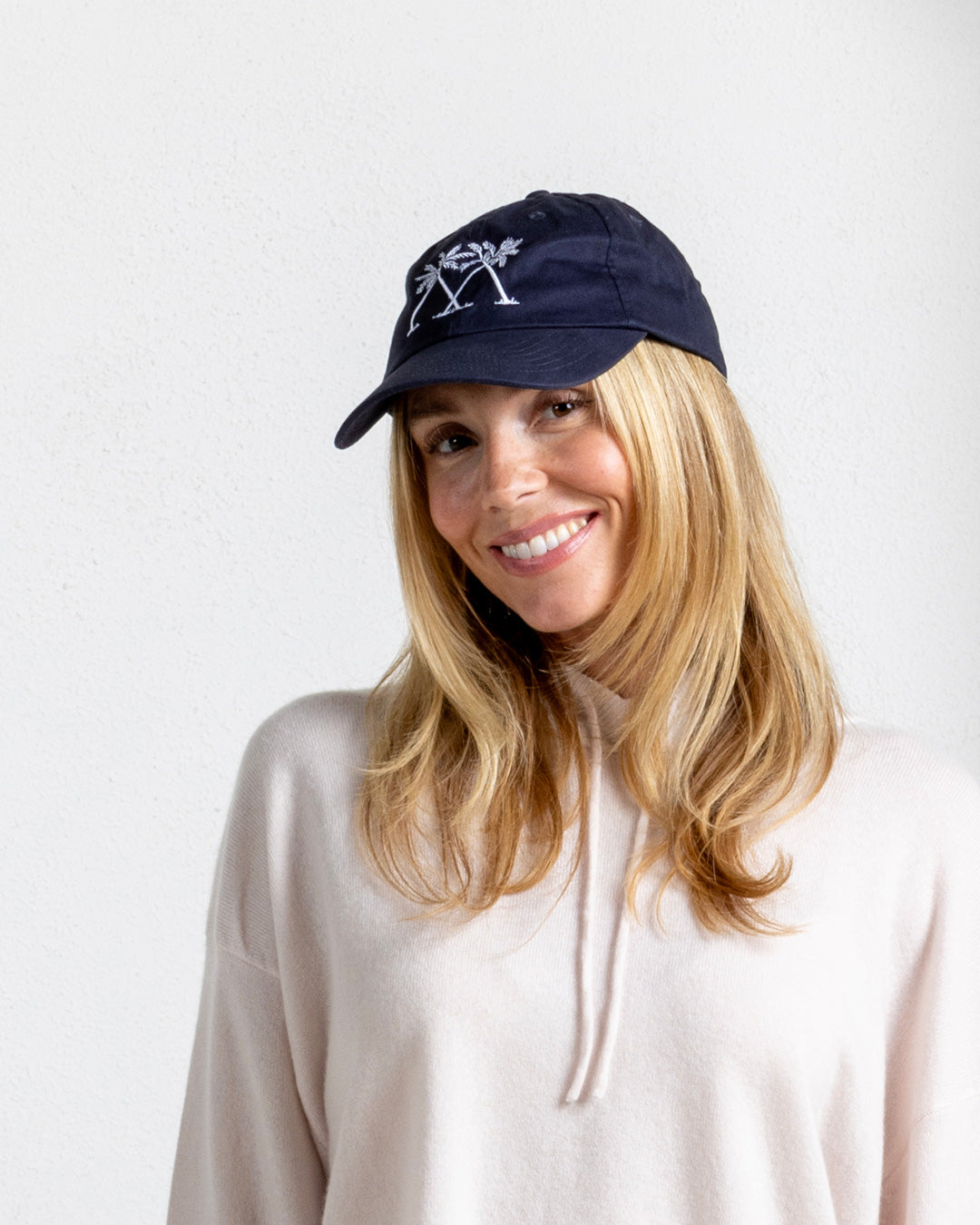 Smiling model in navy baseball cap with white palm logo, Mustique island style paired with luxury cashmere hoodie.