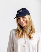 Smiling model in navy baseball cap with white palm logo, Mustique island style paired with luxury cashmere hoodie.