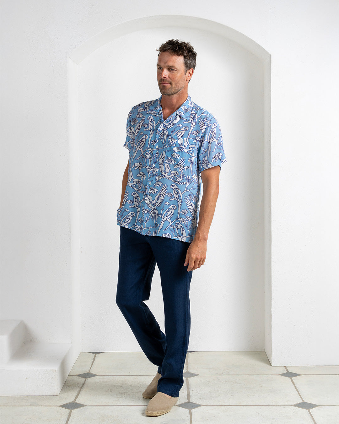 Joss wearing blue silk festival shirt with white parrot print, styled with navy linen trousers – Pink House Mustique resortwear