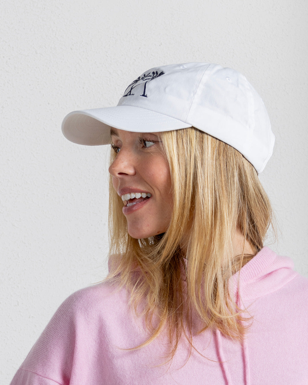 White Mustique palm tree cap styled with pastel hoodie, worn by woman smiling
