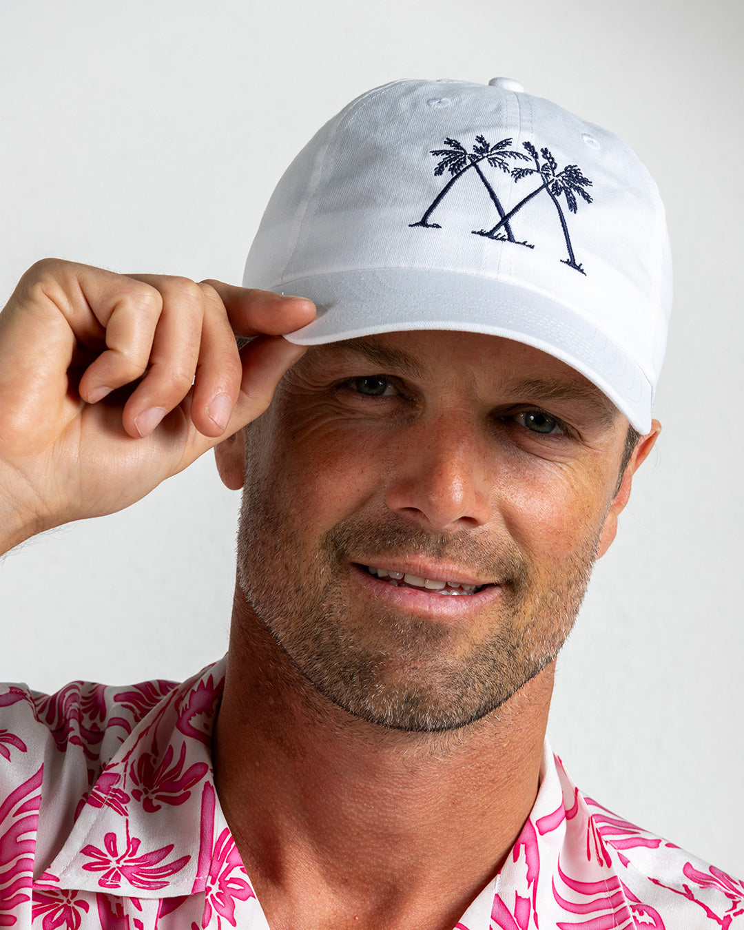 White Mustique Palms logo cap worn by man adjusting brim, pink silk shirt