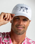 White Mustique Palms logo cap worn by man adjusting brim, pink silk shirt