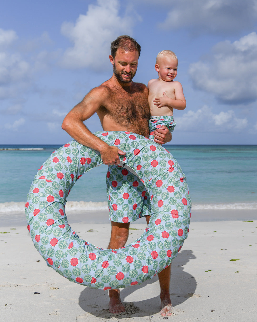 Father and toddler in matching Watermelon print swim shorts with beach ring on a Caribbean beach