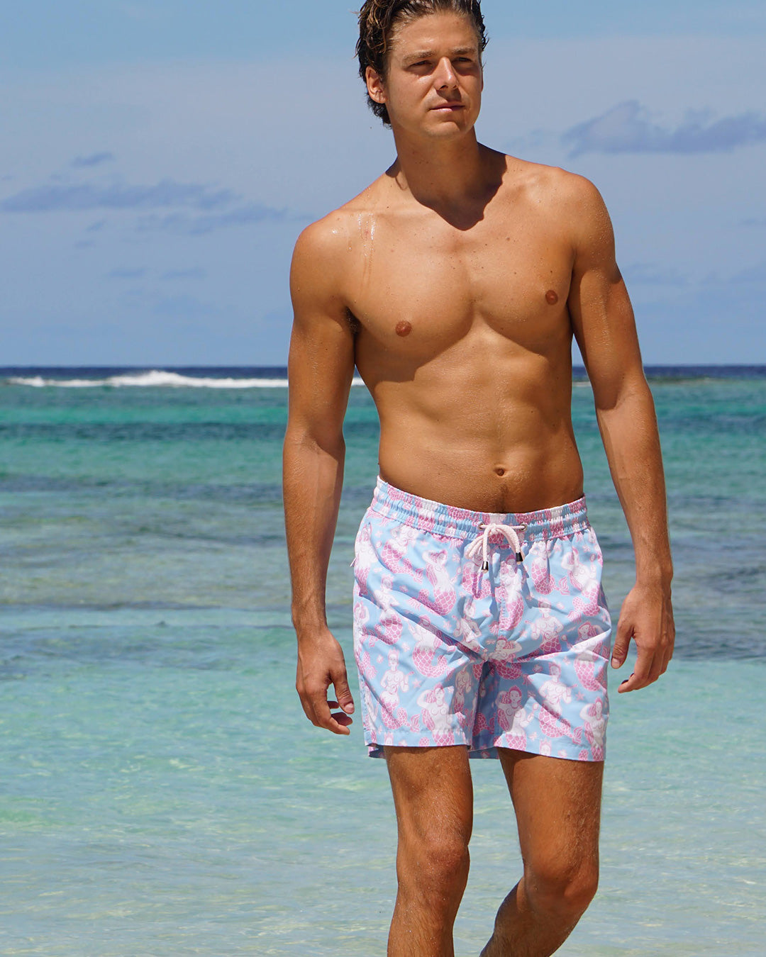 Man wearing Pink House Mustique Mermaid Pink Blue swim shorts walking in the sea at Lagoon Bay Mustique