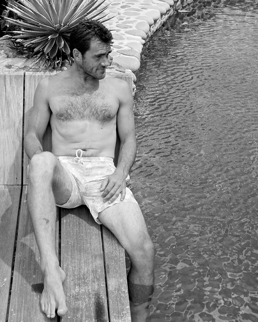 Black and white image of a man relaxing poolside in Mermaid Pink Blue swim shorts in Mustique