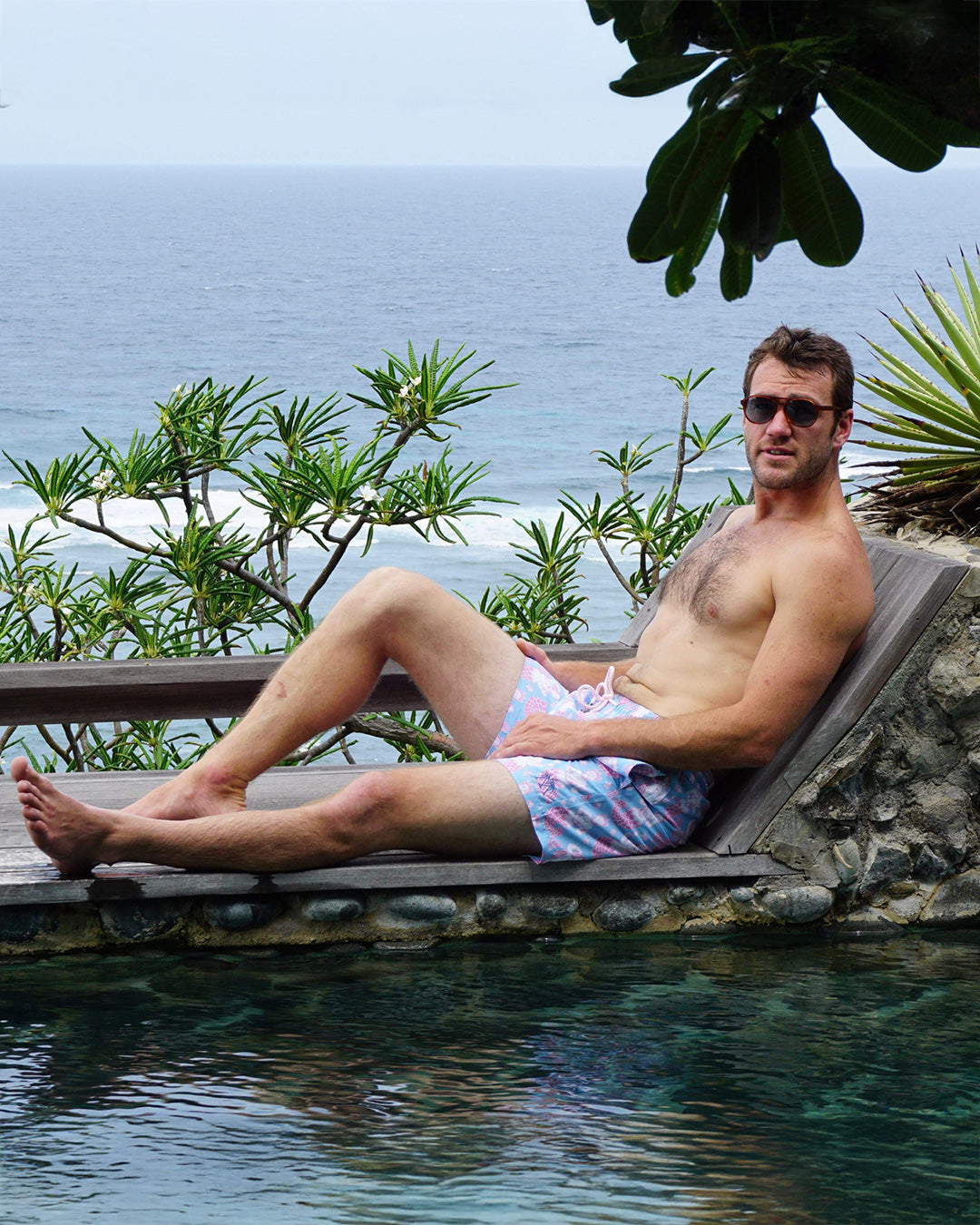 Man relaxing poolside in Pink House Mustique Mermaid Pink Blue swim shorts overlooking the Caribbean Sea