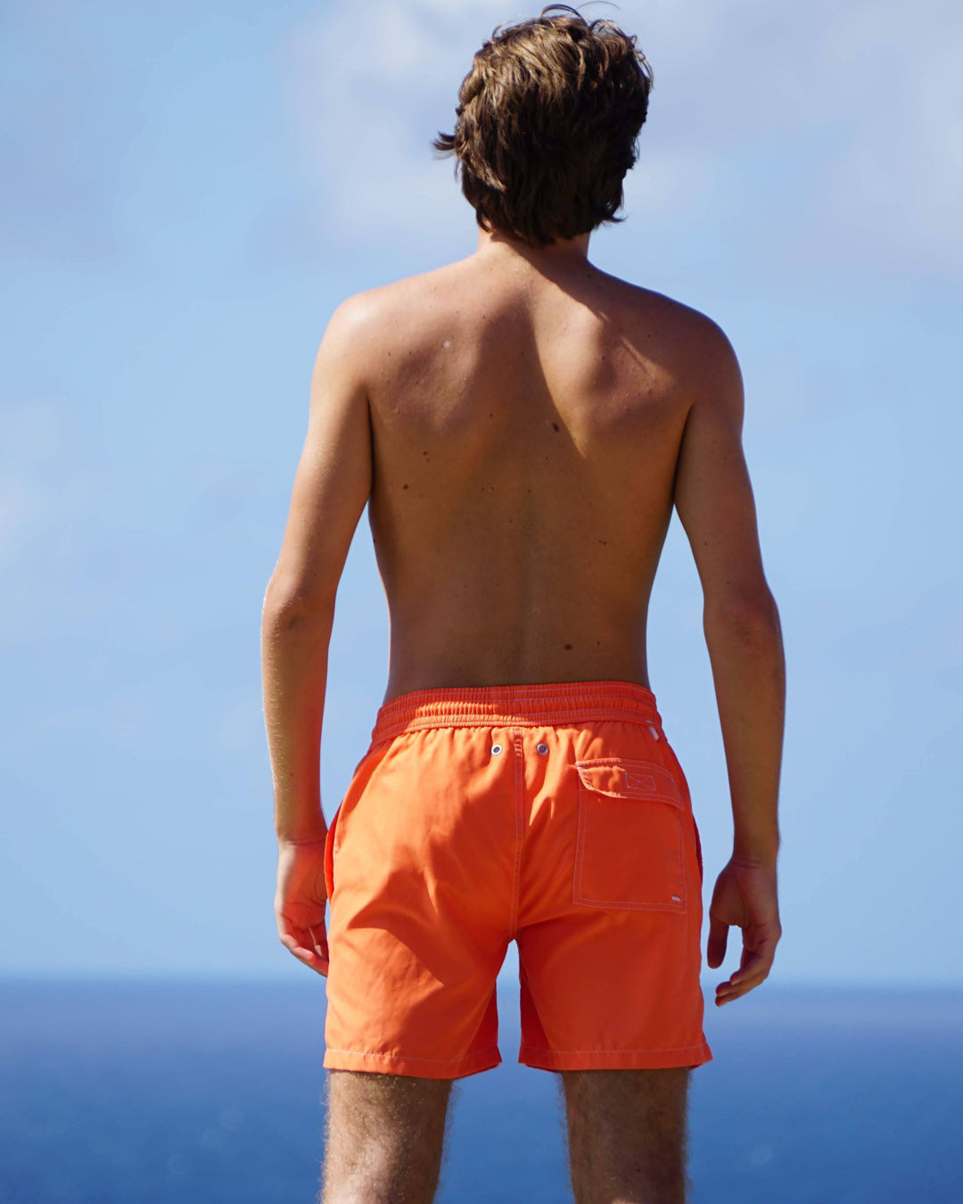 Men’s orange swim shorts back view by ocean, classic beach style and tailored fit