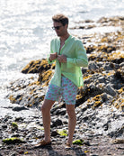 Mens pistachio green linen shirt with collarless grandad Nehru collar, buttoning on beach with green pink swim shorts