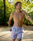 Man wearing pink swim shorts with a pattern, standing on a sandy beach with greenery in the background.