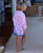 Child wearing a pink floral linen shirt and blue shorts walking on a tiled floor.
