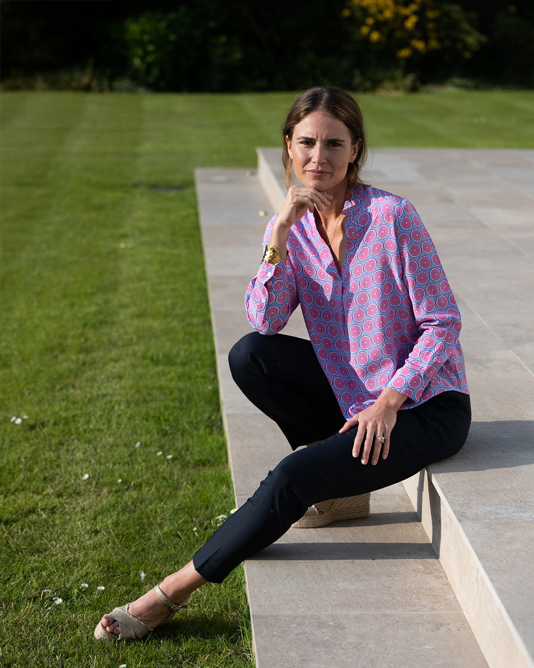 Woman sitting on a stone bench outdoors wearing a pink patterned shirt and black pants.