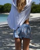 Rear view of model in blue Parrot print silk shorts on a Caribbean beach under flowing linen shirt