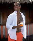 Man wearing Pink House Mustique Optic White Linen Shirt – lightweight breathable linen shirt styled open with vibrant orange tailored swim shorts, perfect for luxury beachwear and tropical resort style
