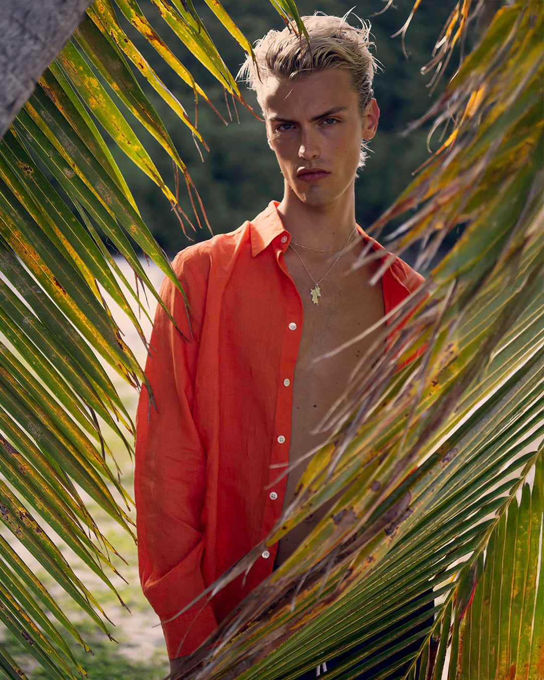 Model wearing Pink House men’s pure linen shirt in orange, styled open at the beach with palm leaves and Caribbean backdrop