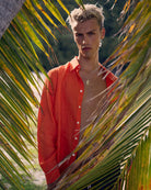 Model wearing Pink House men’s pure linen shirt in orange, styled open at the beach with palm leaves and Caribbean backdrop