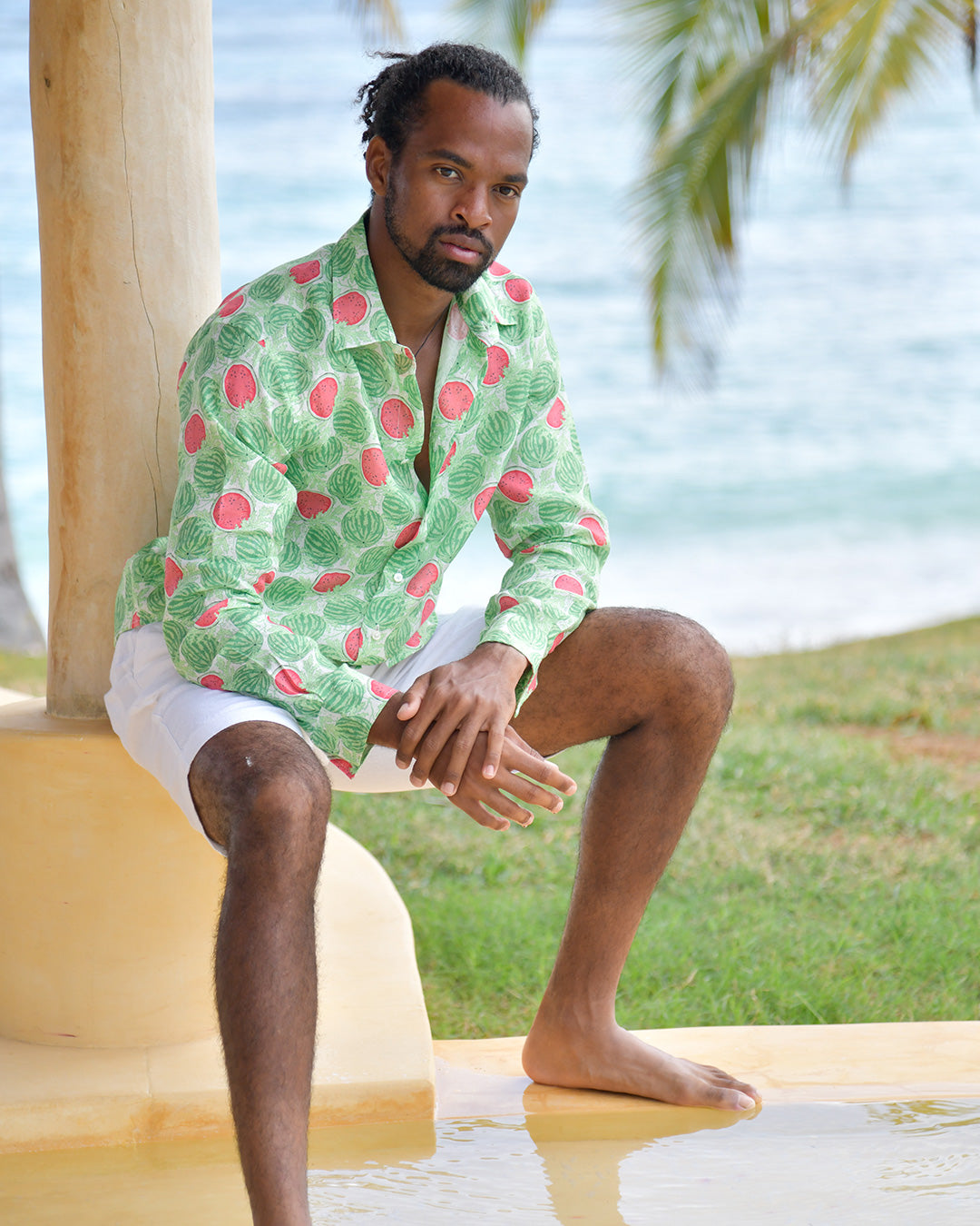 Man seated in Watermelon Green linen shirt with white shorts beside the sea in an island setting
