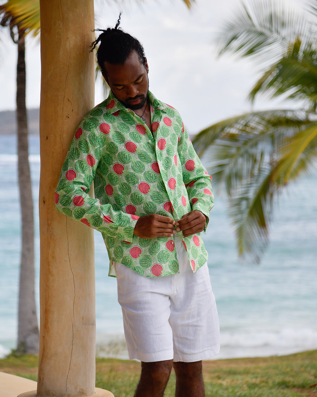 Man buttoning Watermelon Green linen shirt with white shorts in a relaxed tropical island setting