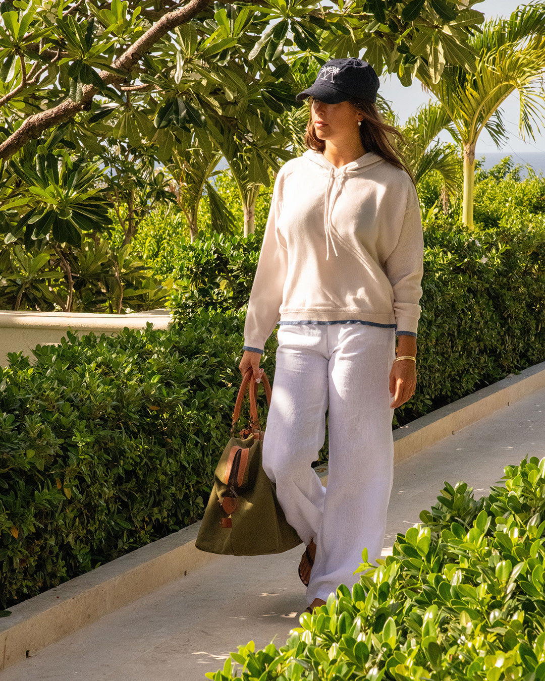 Woman wearing a beige 100% Mongolian cashmere hoodie styled with white linen Flora trousers and navy cap by Pink House Mustique